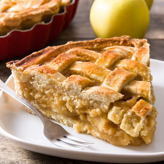 White plate with slice of apple pie in foreground. Background of apple pie and apple.